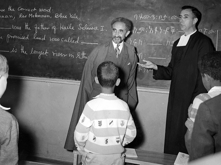 Emperor Haile Selassie of Ethiopia stands in front of a chalk board looking out at a class of students.  Next to him, introducing him, is a Christian Brother.